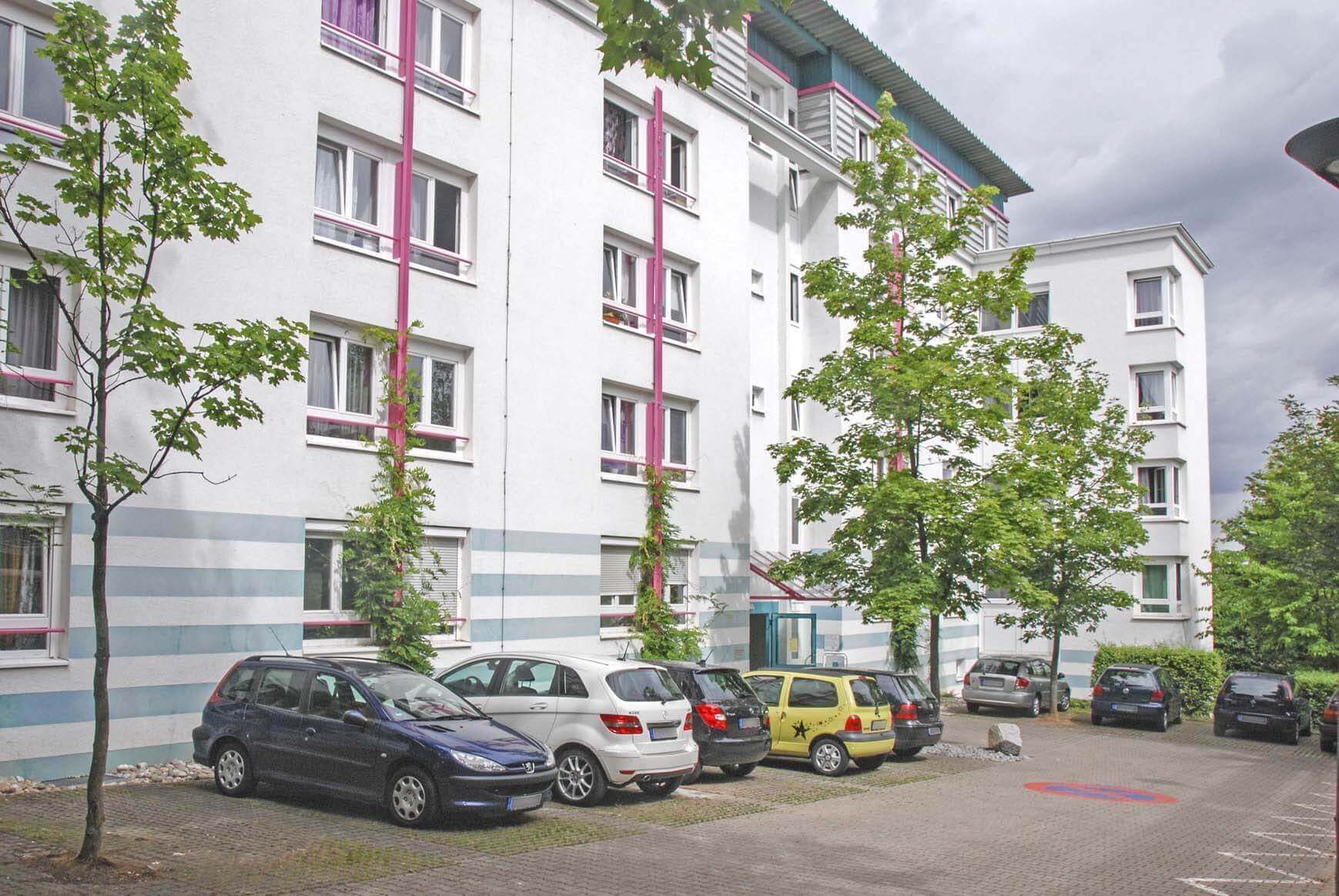 Side view of a bright residential building with blue and pink color accents and several parking spaces in front of it.