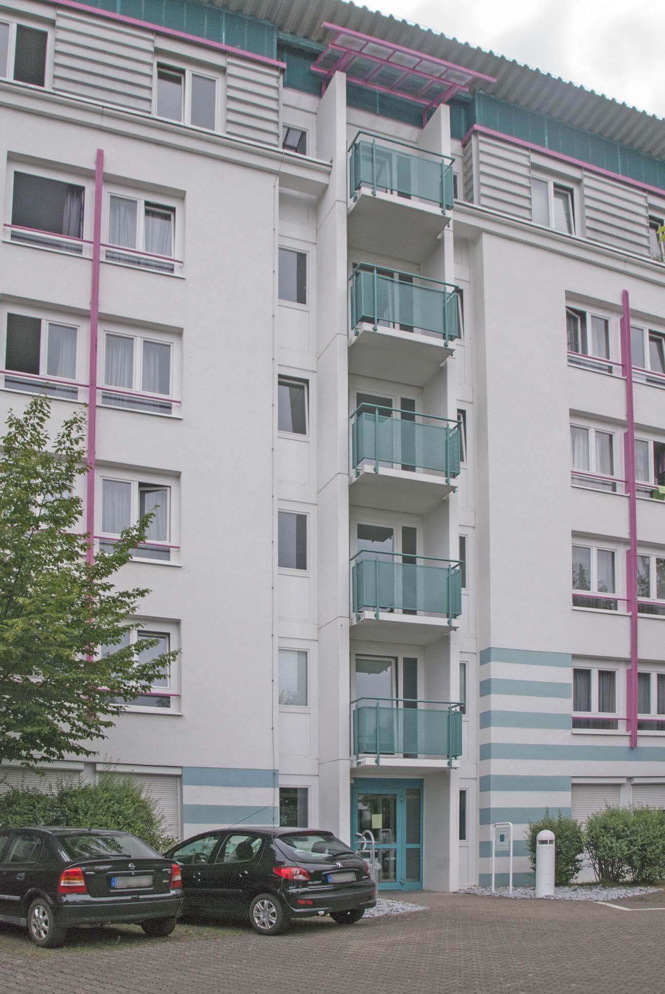 Entrance of a multi-storey building with blue frame, balconies and parked cars in front of it.