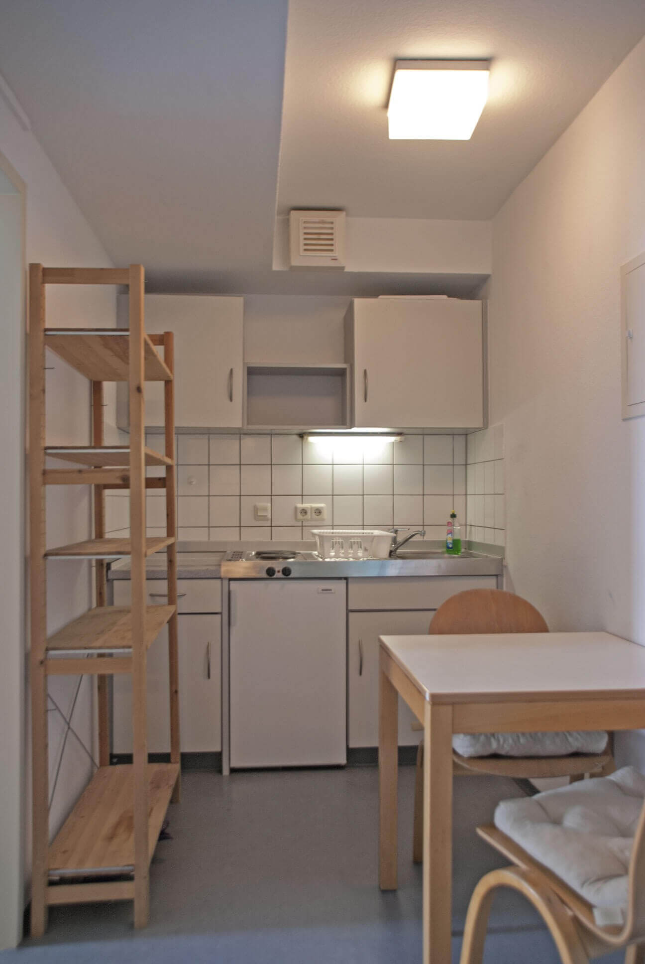 Kitchenette with sink, stove, wall cupboards and a small shelf. Table with chair in the foreground.