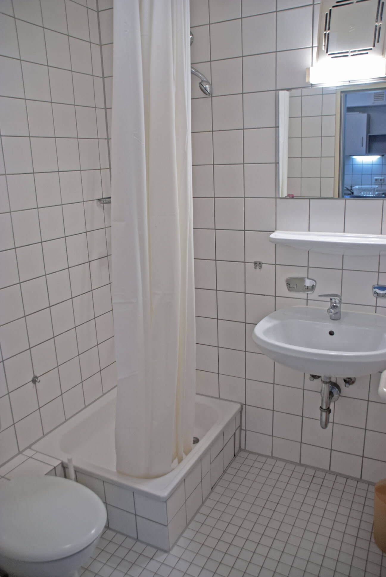 Small bathroom with shower tray, curtain, washbasin and toilet. White tiles on walls and floor.
