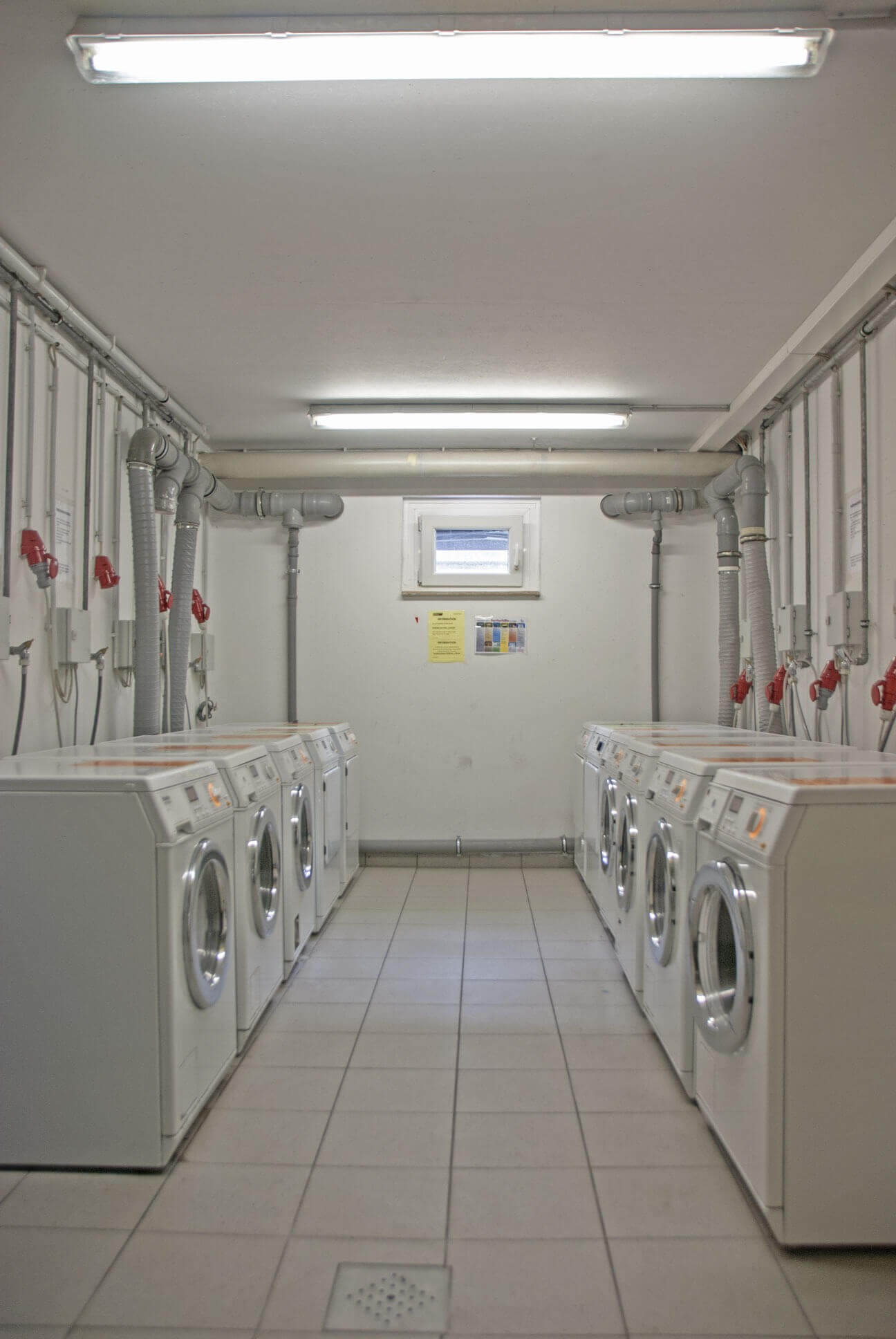 Laundry room with two rows of washing machines and numerous electricity and water connections on the walls.