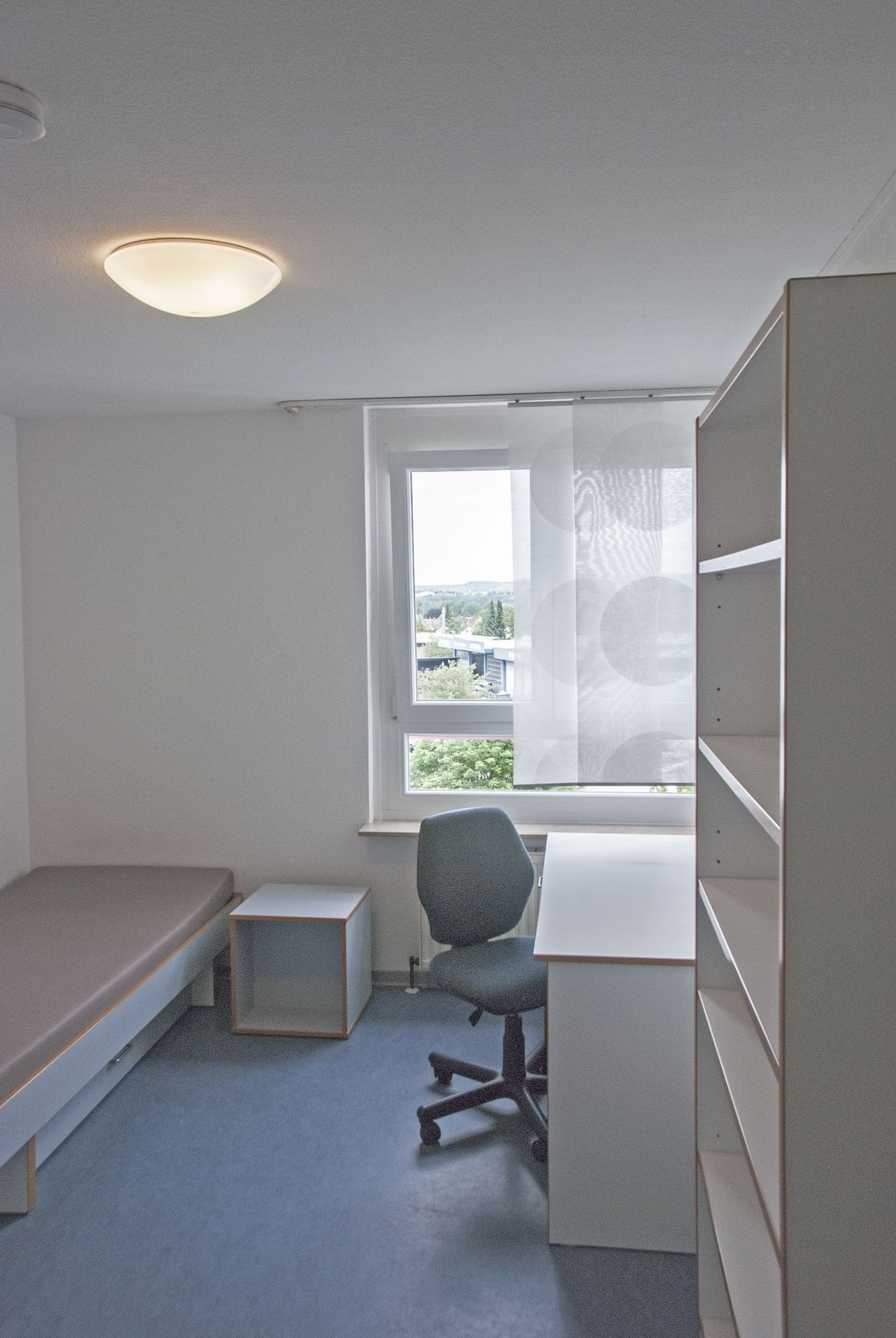 Simply furnished single room with bed, desk, chair and shelf. Window with a view of the surroundings.