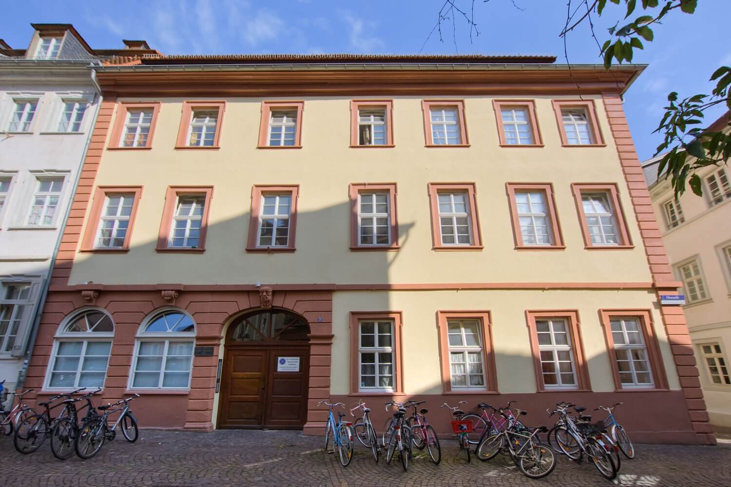 Außenansicht eines dreistöckigen Altbaus mit gelber Fassade, roten Fensterrahmen und davor geparkten Fahrrädern.