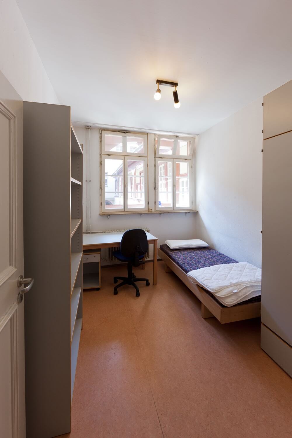 Einzelzimmer mit Bett, Schreibtisch, Regal und großem Fenster mit Blick auf den Laubengang.