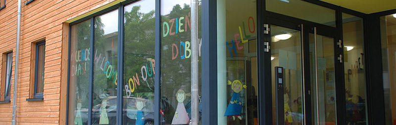 Blick auf den Eingang einer Kita mit großen Fenstern. In den Scheiben sind bunte Begrüßungen und Kinderzeichnungen zu sehen.