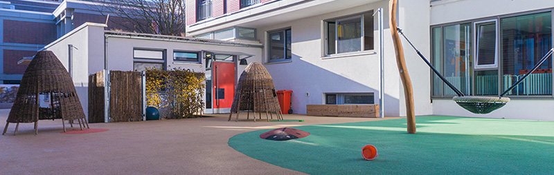Ein leerer, moderner Spielplatz vor einem Kita-Gebäude. Die Fläche ist mit Fallschutzboden ausgestattet, Spielgeräte und Weidenhütten stehen bereit.