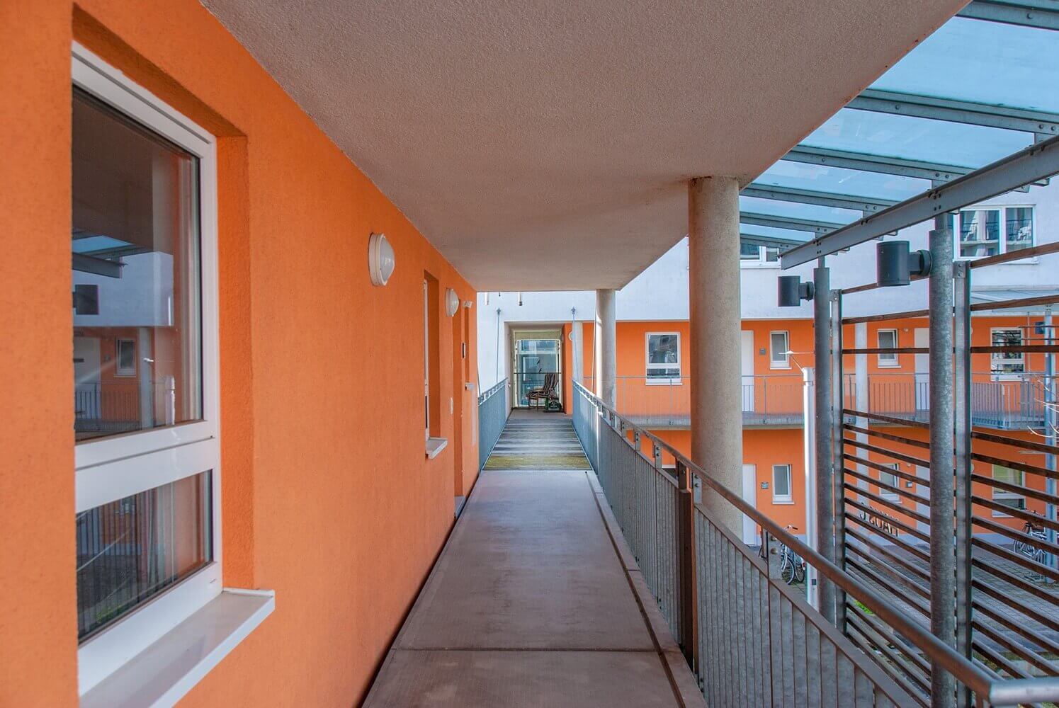 Covered entrance with a view of the open gallery of a student residence, orange-painted façade and glass roofing