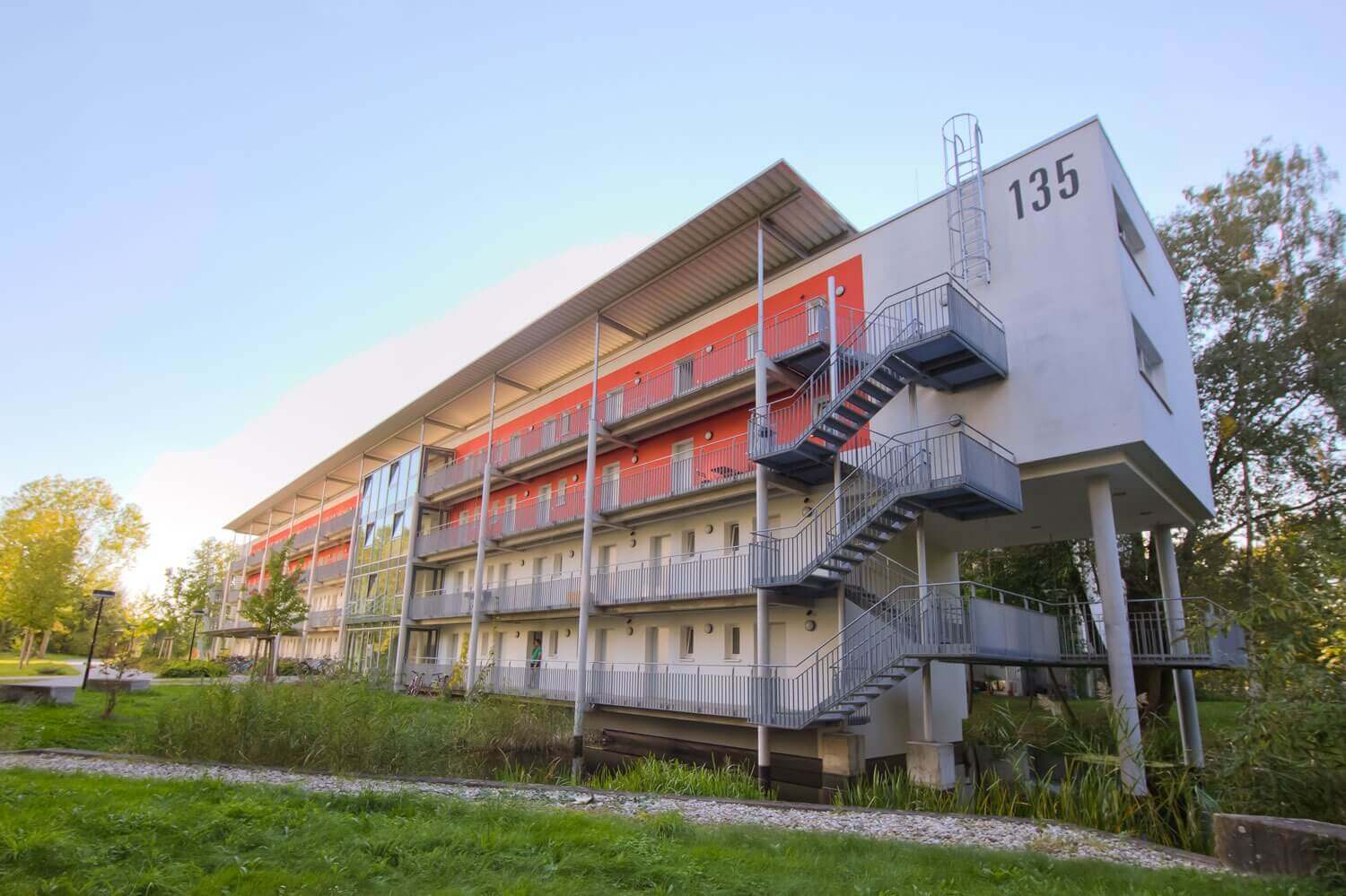 Langgestrecktes, modernes Studentenwohnheim mit auffälliger Außentreppe, roter Fassade und großen Glasfronten inmitten einer begrünten Umgebung