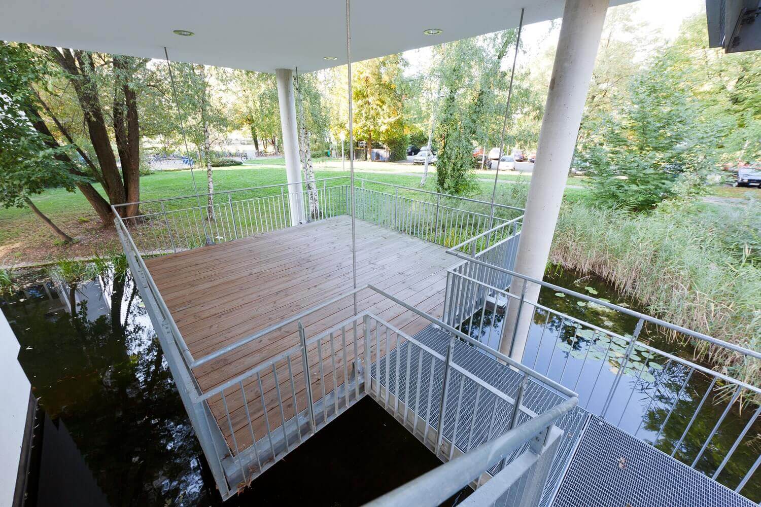 Schwebende, überdachte Holzterrasse mit Metallgeländer über einem Teich, umgeben von Bäumen und einer weitläufigen Grünfläche