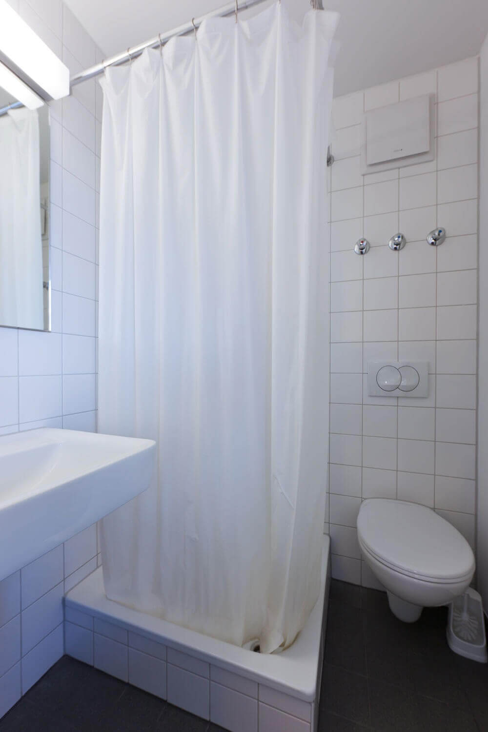 Badezimmer mit weiß gefliester Dusche mit Duschvorhang, WC und Waschbecken – funktional und schlicht eingerichtet.