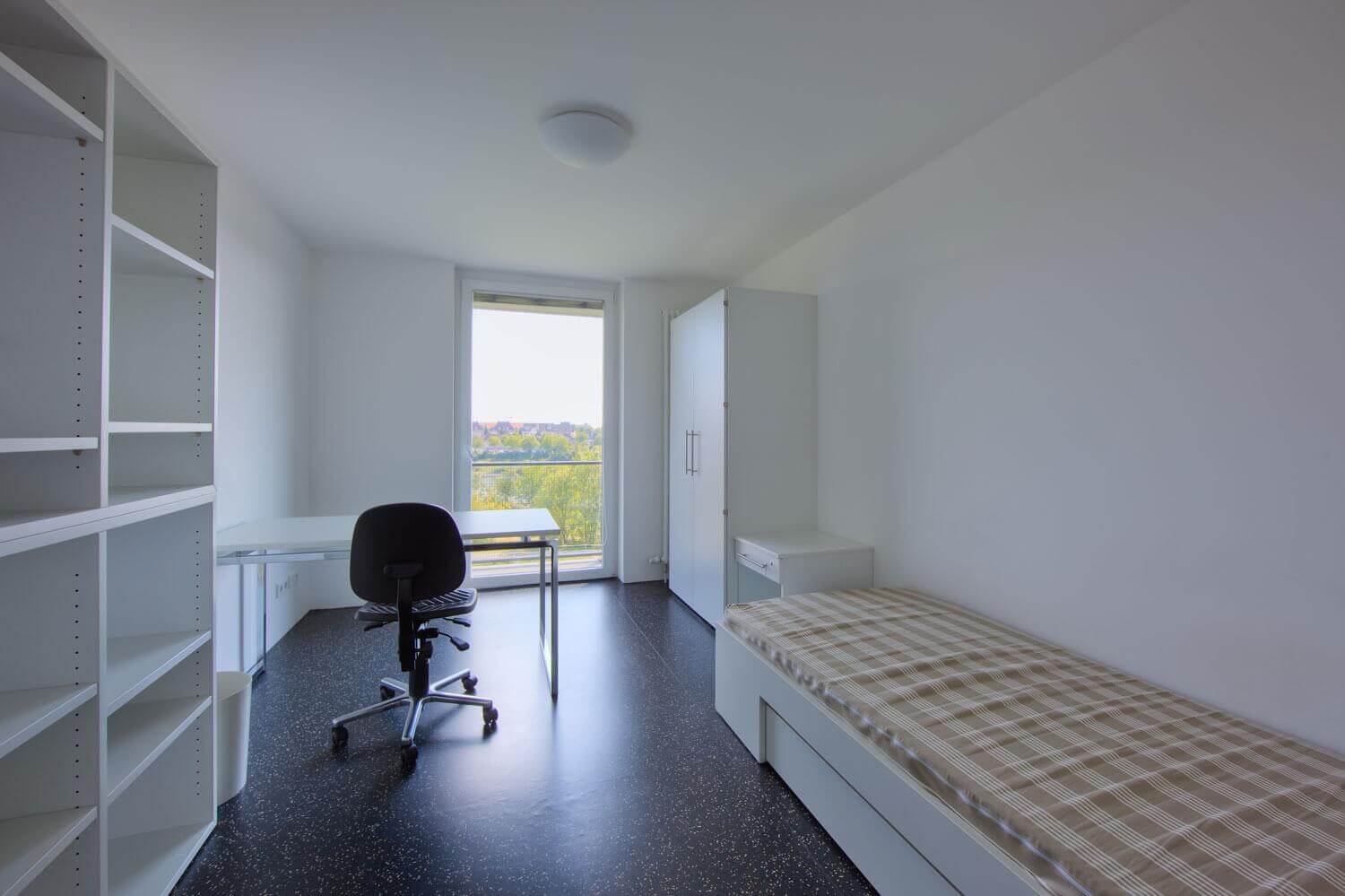 Modern eingerichtetes Einzelzimmer in einem Studierendenwohnheim mit Bett, Schreibtisch, Stuhl, Regal und großem Fenster mit Ausblick ins Grüne.