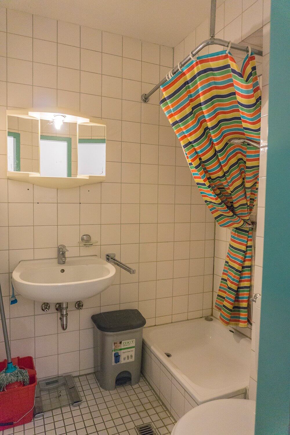 Badezimmer mit weiß gefliester Wand, bunter Duschvorhang, Waschbecken mit Spiegel und kleiner Abfalleimer.