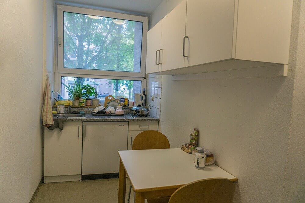 Kleine Gemeinschaftsküche mit Esstisch, zwei Holzstühlen, Oberschränken und einem Fenster mit Blick ins Grüne.