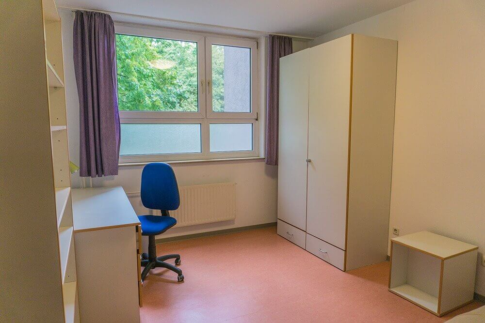 Helles Einzelzimmer im Studentenwohnheim mit großem Fenster, Schreibtisch, Kleiderschrank und hellem Boden.