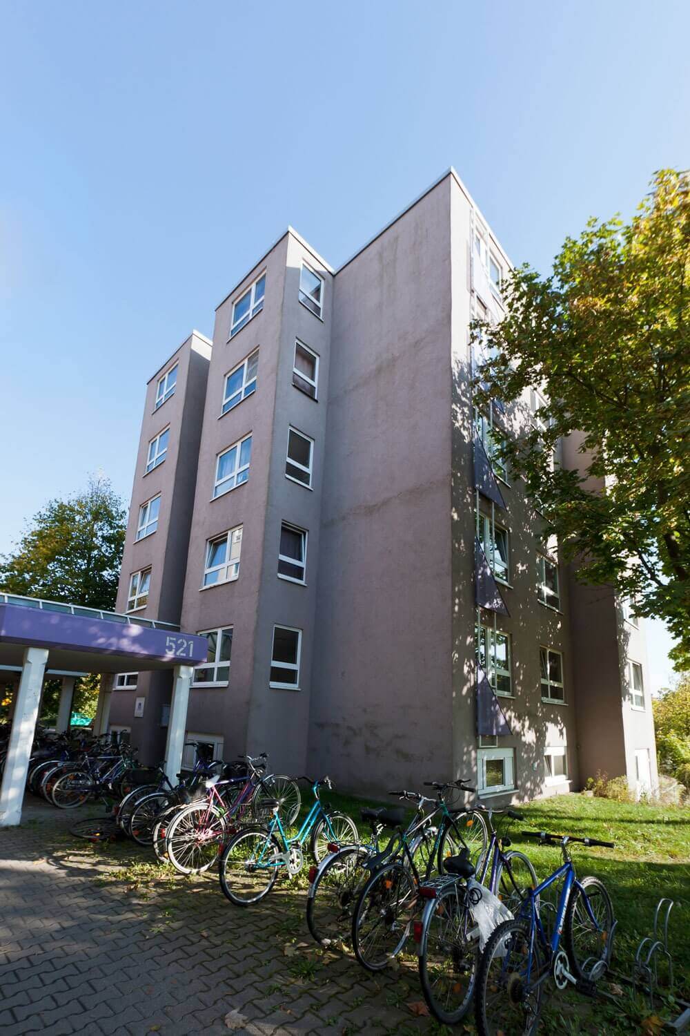 Seitenansicht eines Studentenwohnheims mit grauer Fassade, vielen Fenstern und Fahrradparkplätzen vor dem Gebäude.