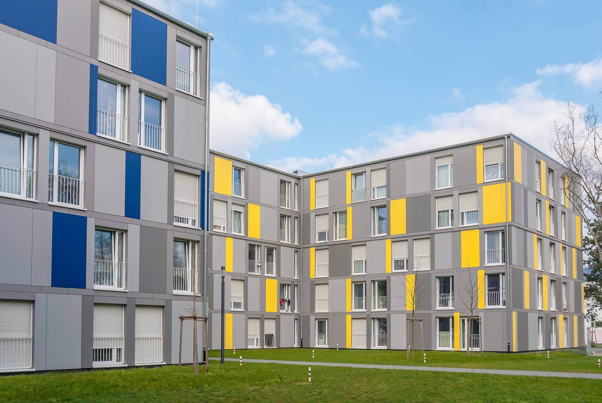 Zwei moderne Wohngebäude mit grauer Fassade und farbigen Akzenten in Blau und Gelb, eingebettet in eine grüne Außenanlage unter blauem Himmel.