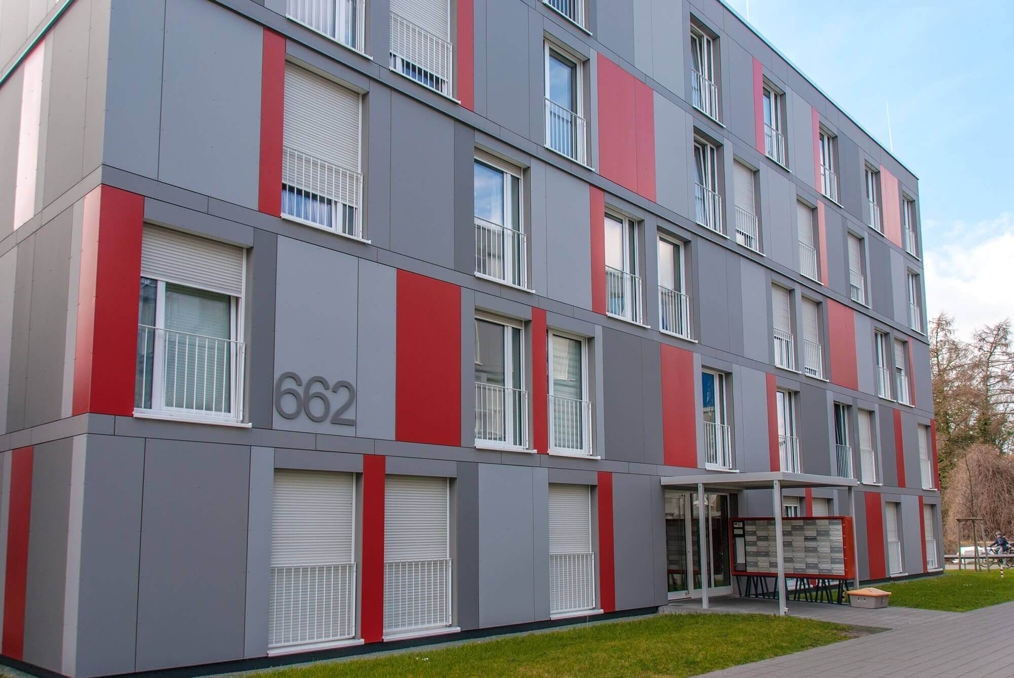Außenansicht eines modernen Wohngebäudes mit grauer und roter Fassade, weißen Fensterrahmen und der Hausnummer 662, aufgenommen bei Tageslicht mit Blick auf den Eingangsbereich.