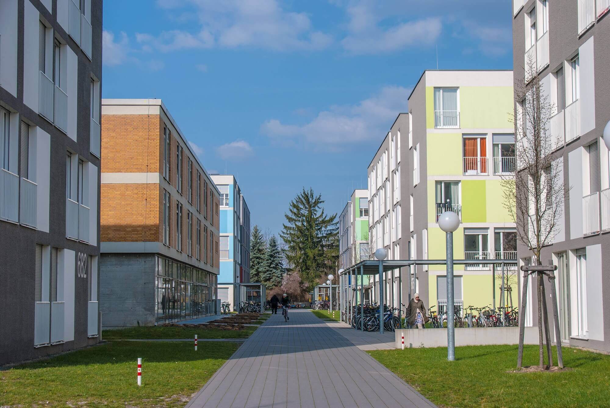 Moderner Innenhof mit mehreren mehrstöckigen Wohnhäusern in bunten Farben wie Grün, Blau und Grau; breite gepflasterte Wege führen zwischen den Gebäuden entlang, flankiert von Rasenflächen, Laternen und zahlreichen Fahrradständern.