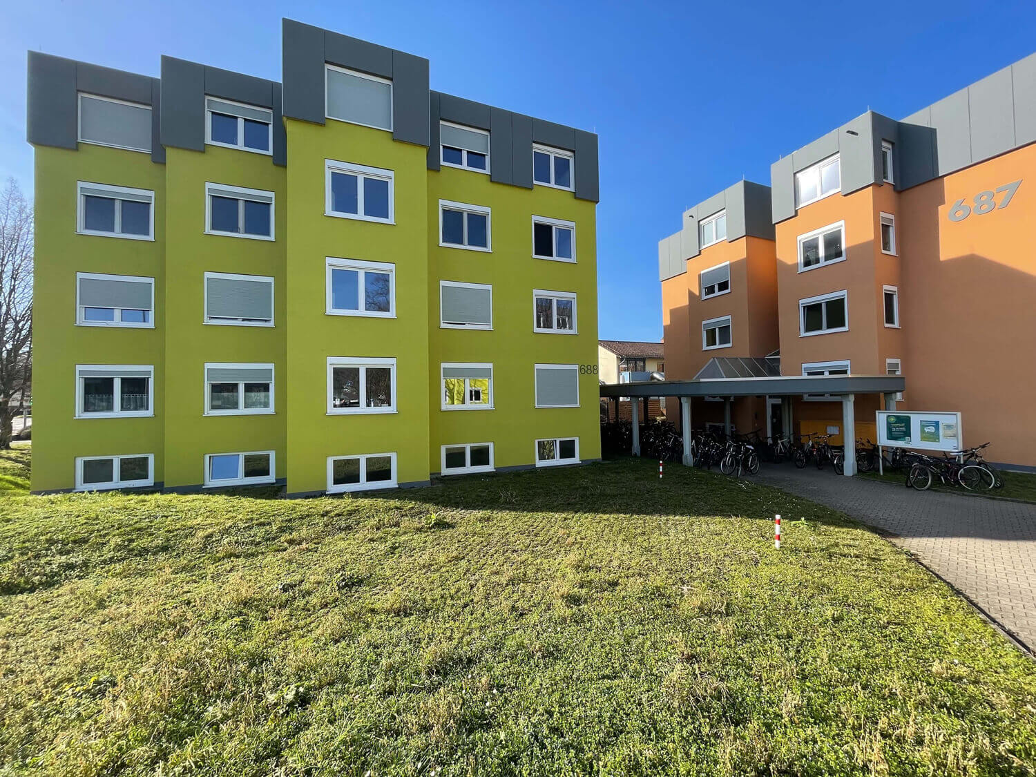 Moderne Wohngebäude mit den Hausnummern 687 und 688 in kräftigem Grün und Orange, davor ein überdachter Fahrradstellplatz und gepflegter Rasenbereich.