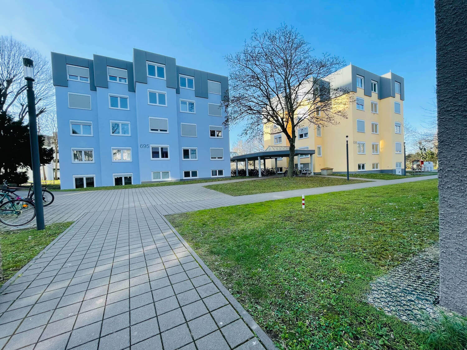Zwei farbenfrohe Wohnheime mit der Hausnummer 695 und 696, umgeben von Rasenflächen und gepflasterten Wegen, unter einem klaren blauen Himmel.