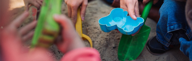 Kinderhände greifen nach buntem Sandspielzeug im Sandkasten. Im Fokus liegt eine blaue Form in Bärenform, daneben grüne Schaufeln.
