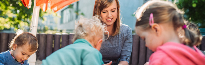 Mehrere Kleinkinder spielen gemeinsam im Sandkasten. Eine Frau betreut die Kinder und beobachtet das Spiel unter einem Sonnensegel.