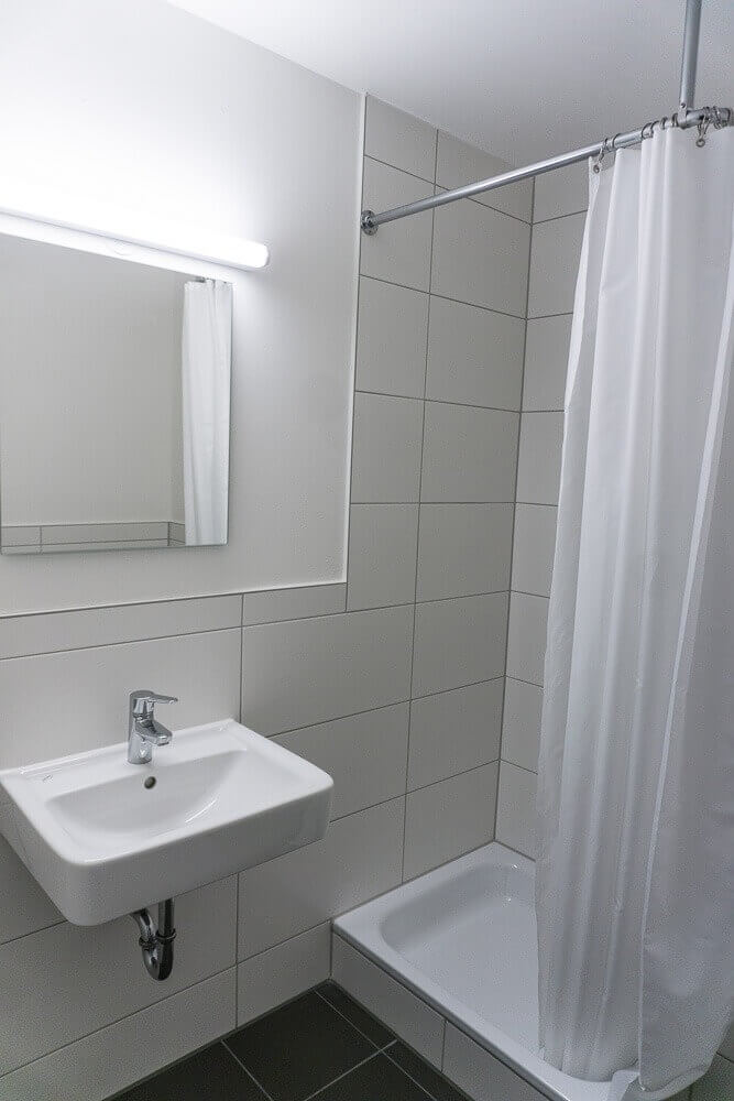 Weiß gefliestes Badezimmer mit Waschbecken, Spiegel, Dusche mit Vorhang und wandhängendem WC in funktionaler, moderner Ausstattung.