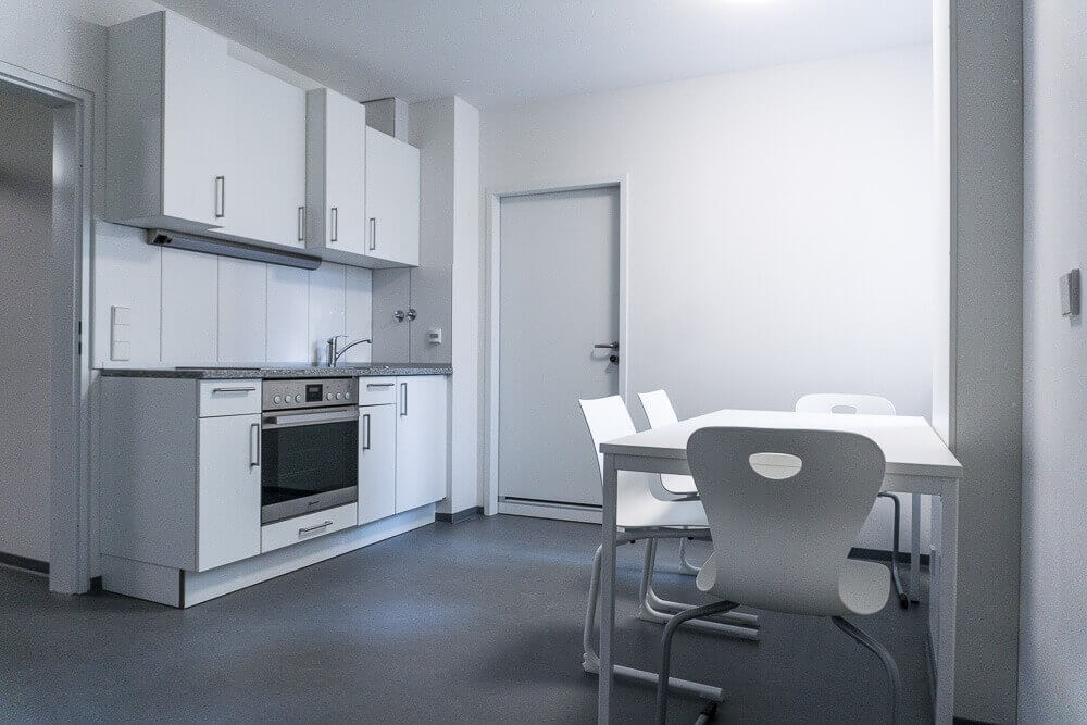 Gemeinschaftsküche mit Esstisch und vier weißen Stühlen, moderner Küchenzeile inklusive Herd, Spüle und Oberschränken in schlichtem Design.