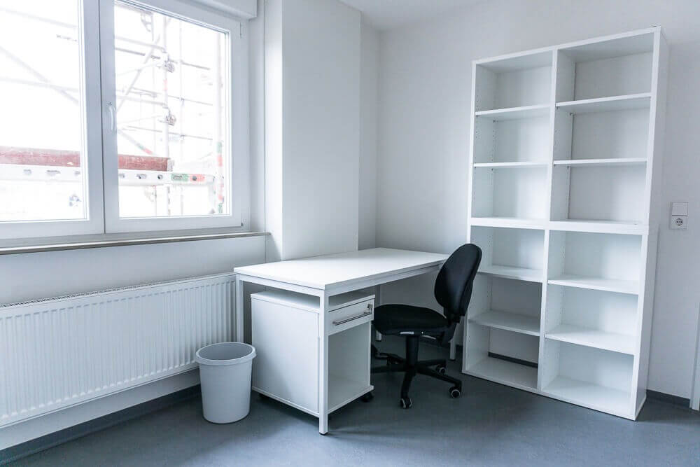 Helles, minimalistisch eingerichtetes Zimmer mit großem Fenster, einem weißen Schreibtisch, Bürostuhl und Regal, ideal zum Lernen und Arbeiten.