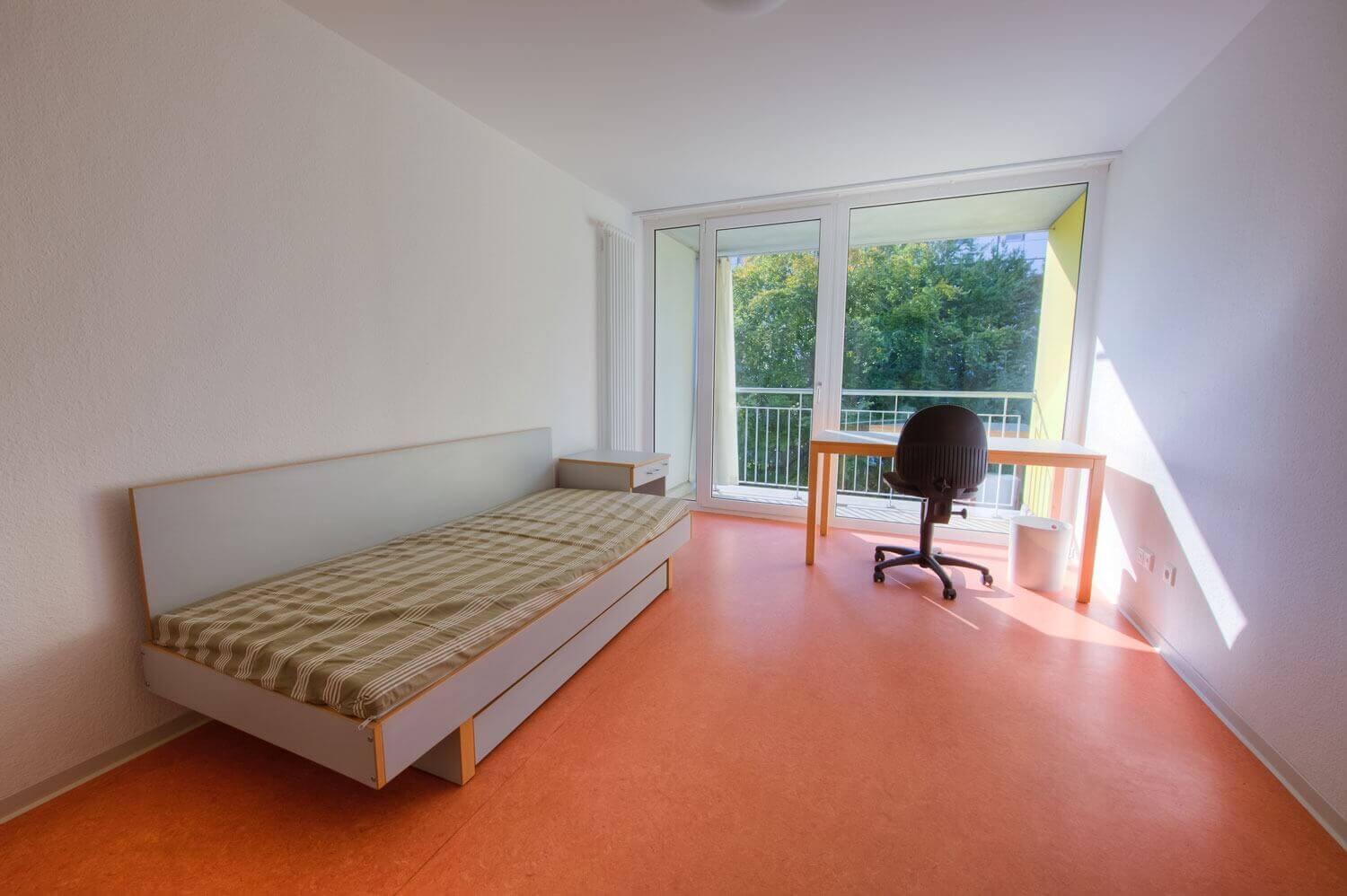 Studentenzimmer mit großem Fenster, Schreibtisch und Bett – heller Raum mit Blick ins Grüne, ideal zum Lernen und Wohnen.