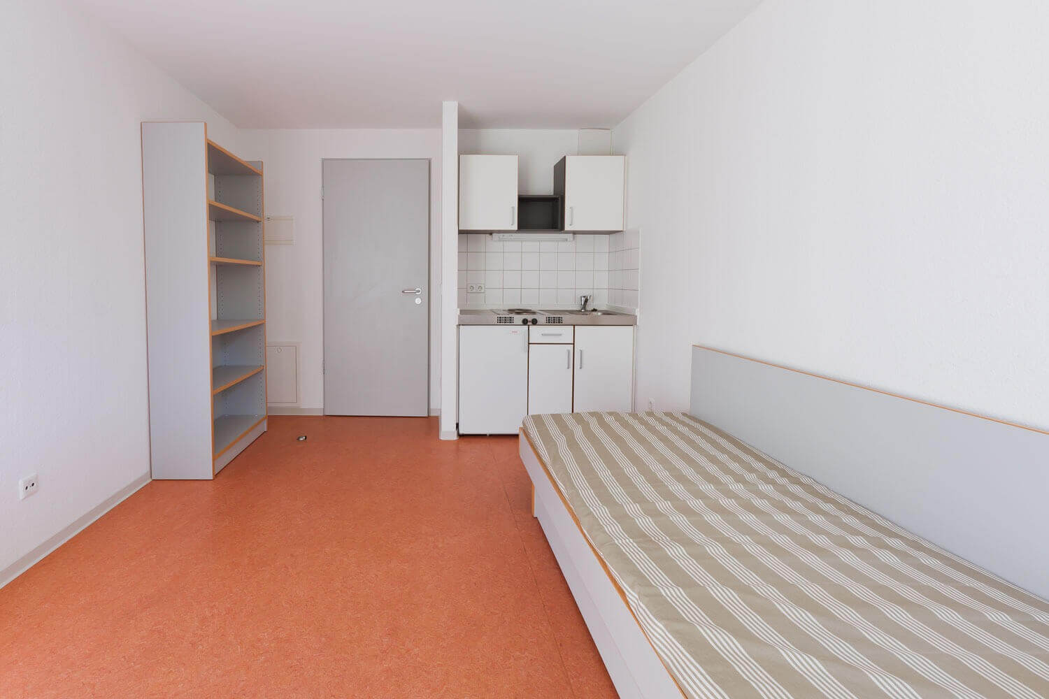 Einzelzimmer im Studentenwohnheim mit Schrank, Regal, Bett und integrierter Pantryküche in heller, minimalistischer Gestaltung.