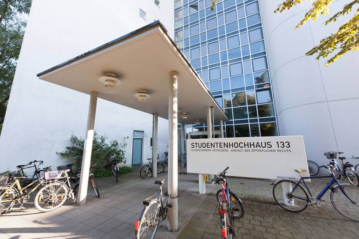 Eingangsbereich eines Studentenwohnheims mit überdachtem Zugang, Fahrradstellplätzen und dem Schild „Studentenhochhaus 133“ des Studierendenwerks Heidelberg.