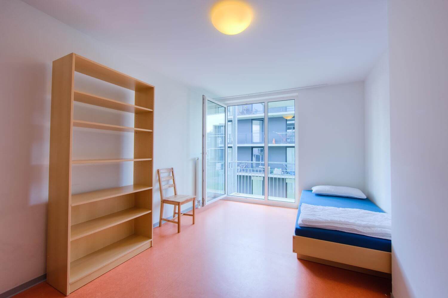 Single room with blue bed, large shelf, chair and floor-to-ceiling window overlooking the inner courtyard