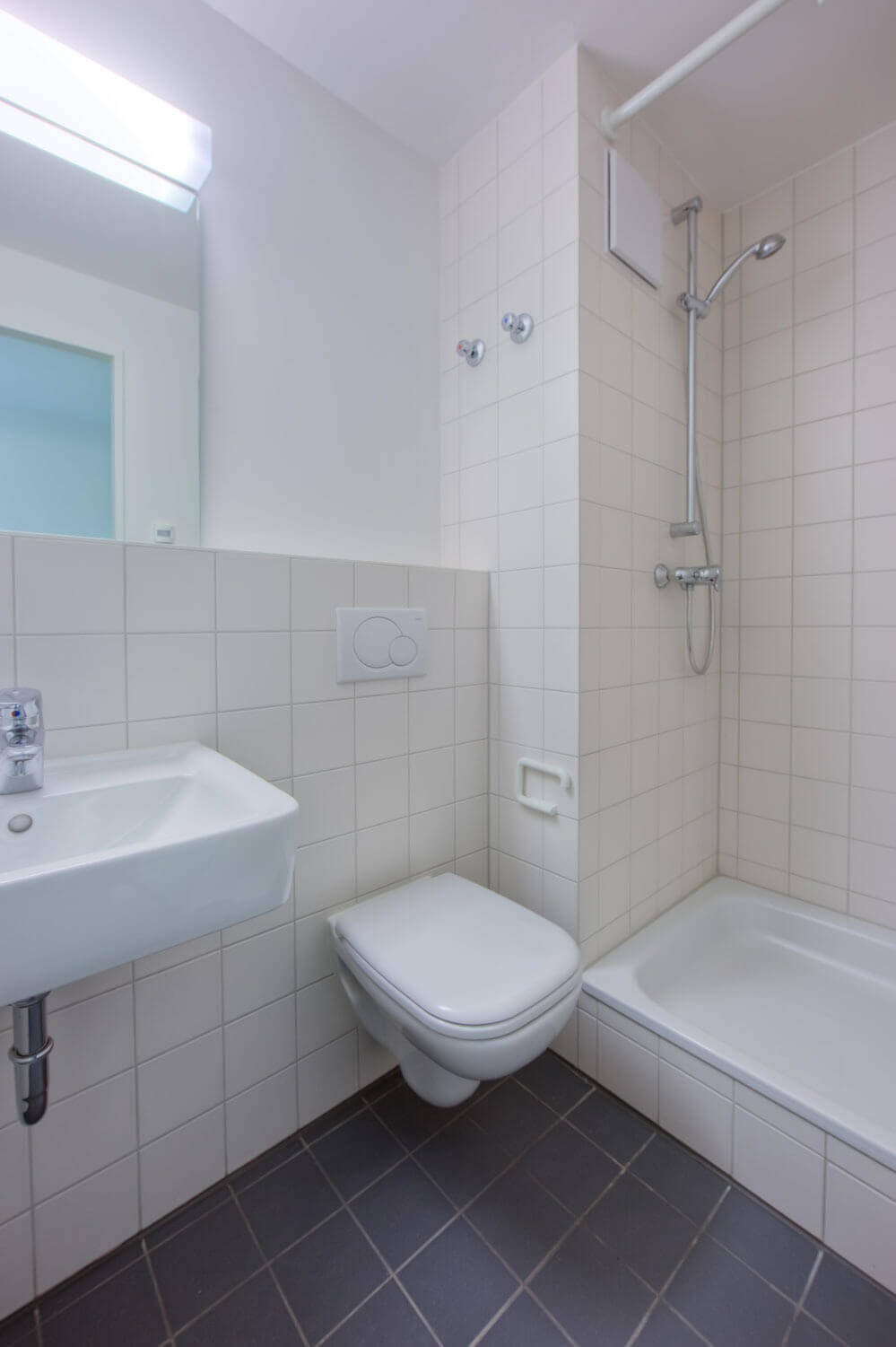 Kompaktes, funktionales Badezimmer mit hellen Wandfliesen, grauem Boden, Toilette, Waschbecken und integrierter Dusche mit Duschvorhangstange; alles in hellen, pflegeleichten Materialien gehalten.