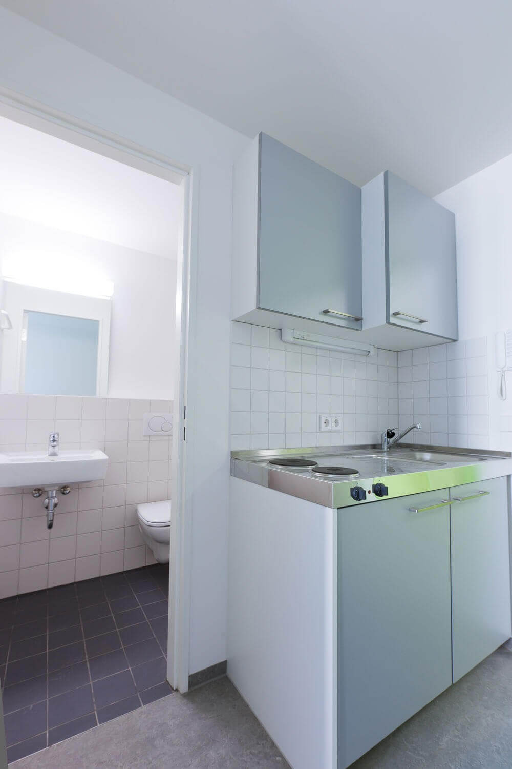Kleine Kochnische mit zwei Herdplatten und Spüle, weißen Fliesenspiegeln und Hängeschränken; im Hintergrund ist das angrenzende Badezimmer mit Waschbecken und Toilette sichtbar.