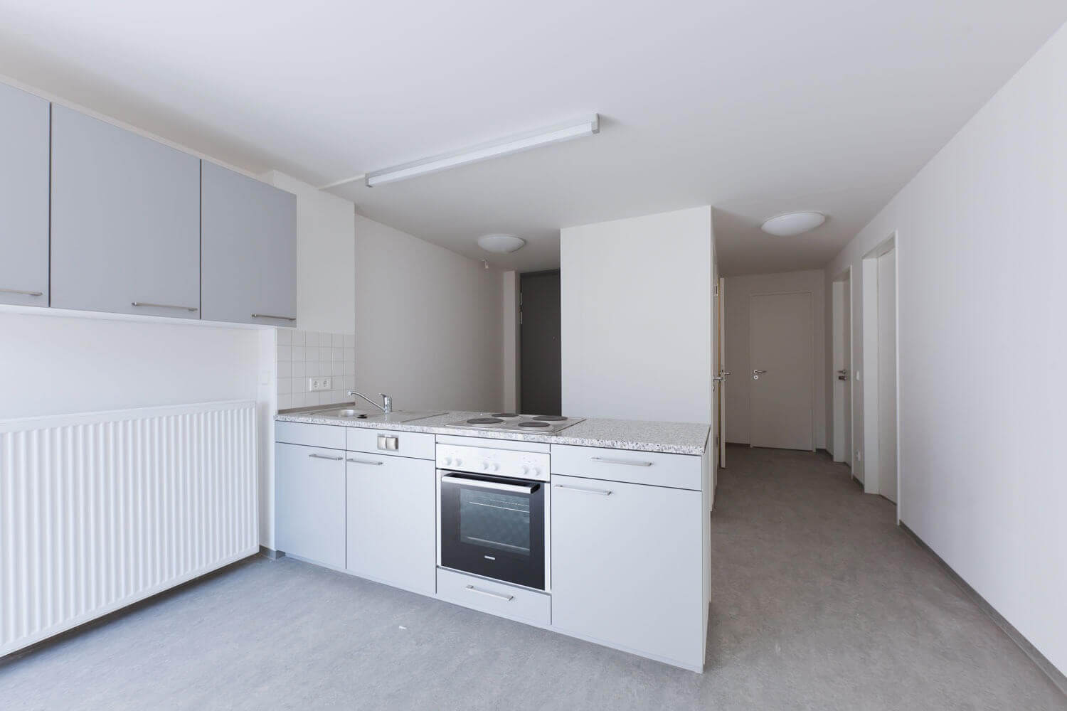 Offene, moderne Küchenzeile in einem Apartment mit hellgrauen Schränken, Elektroherd mit Backofen und Spüle auf einer Arbeitsplatte aus Stein; weiß gestrichene Wände und offener Flur im Hintergrund.