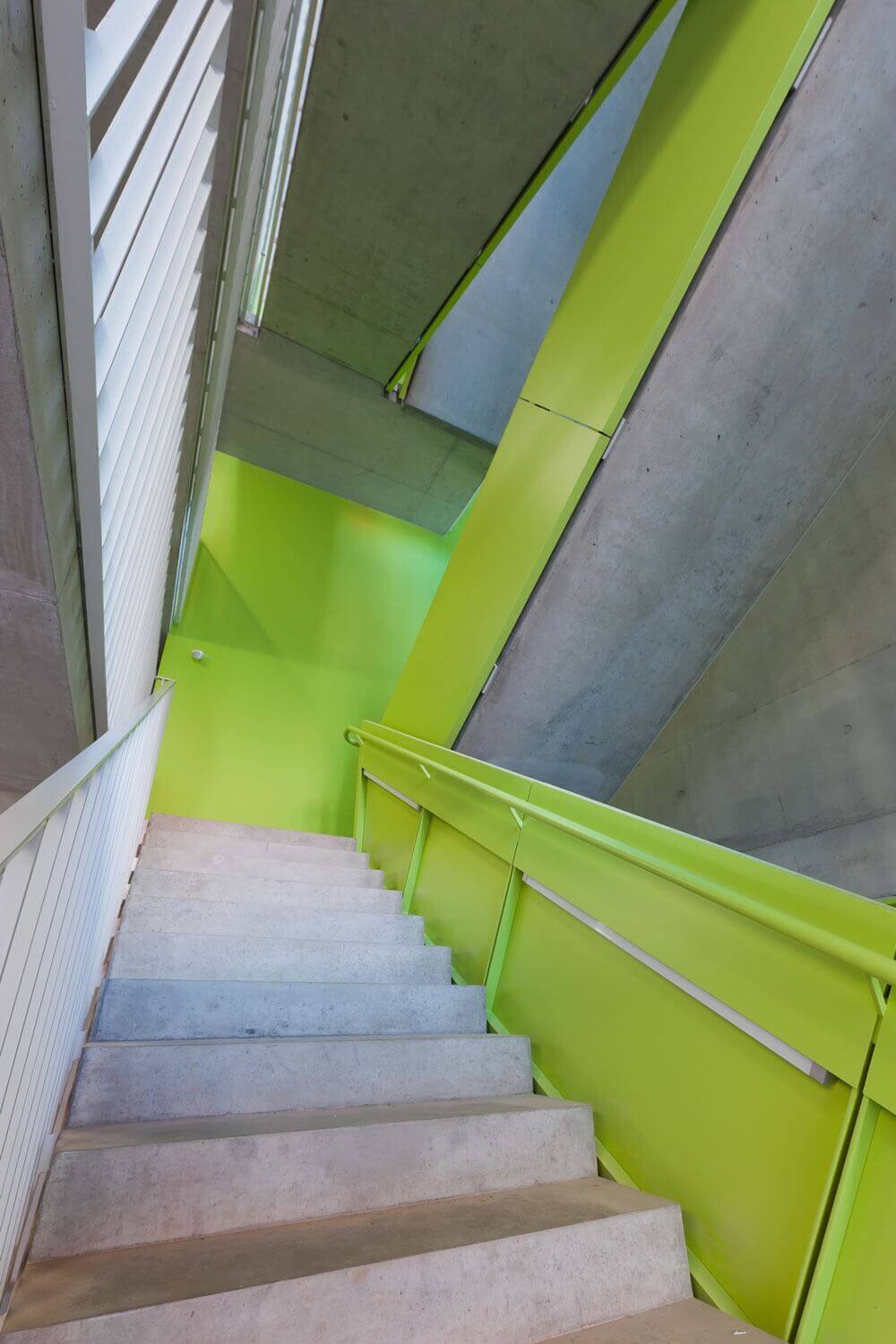 Innenansicht eines auffällig gestalteten Treppenhauses mit signalgrünen Wänden und Geländern sowie Sichtbetonflächen; moderne Architektur mit offenen Treppenläufen und asymmetrischen Perspektiven.
