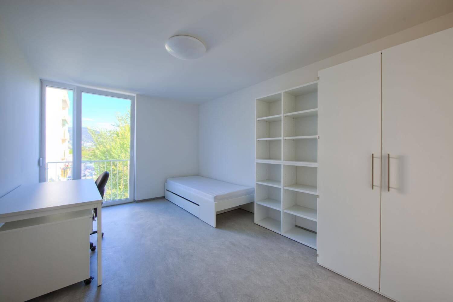 Helles Einzelzimmer mit großen bodentiefen Fenstern, schlichtem weißen Schreibtisch und Drehstuhl, einem weißen Bett mit ausziehbarem Unterbett sowie einem hohen Regal und einem zweitürigen Kleiderschrank.
