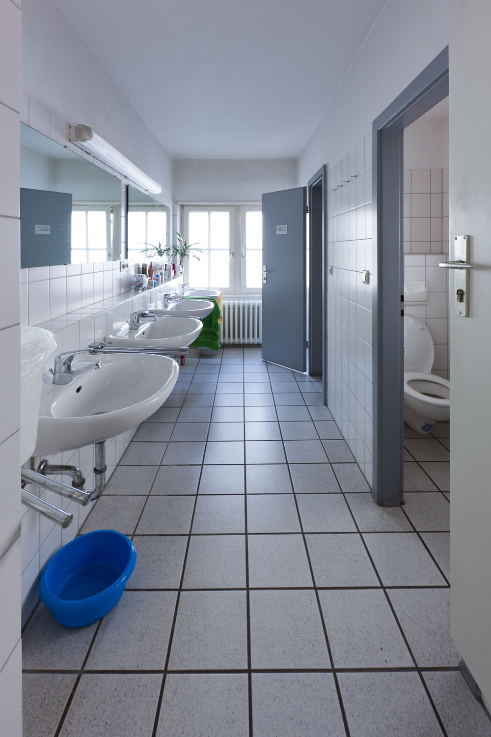 Gemeinschaftsbad mit mehreren weißen Waschbecken entlang einer gefliesten Wand, großem Spiegel, zwei geöffneten WC-Kabinen und Tageslicht durch ein Fenster am Ende des Flurs.