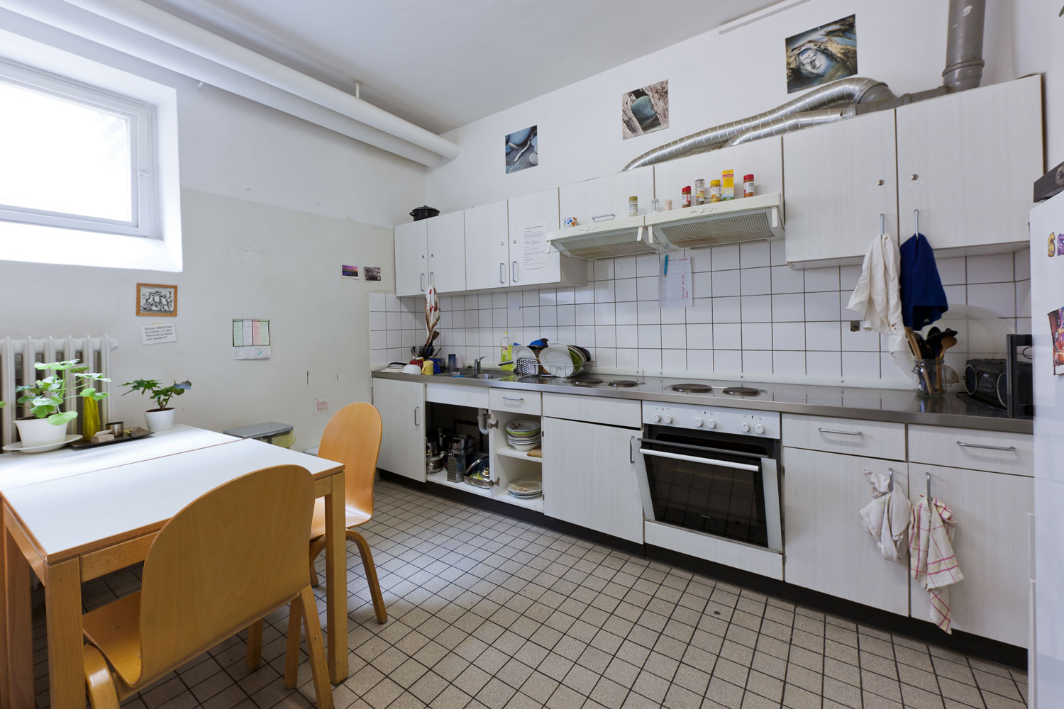 Gemeinschaftsküche mit mehreren Hängeschränken, großem Herd und Cerankochfeld, Edelstahlspüle und Esstisch mit zwei Holzstühlen; persönliche Gegenstände und Pflanzen verleihen eine wohnliche Atmosphäre.