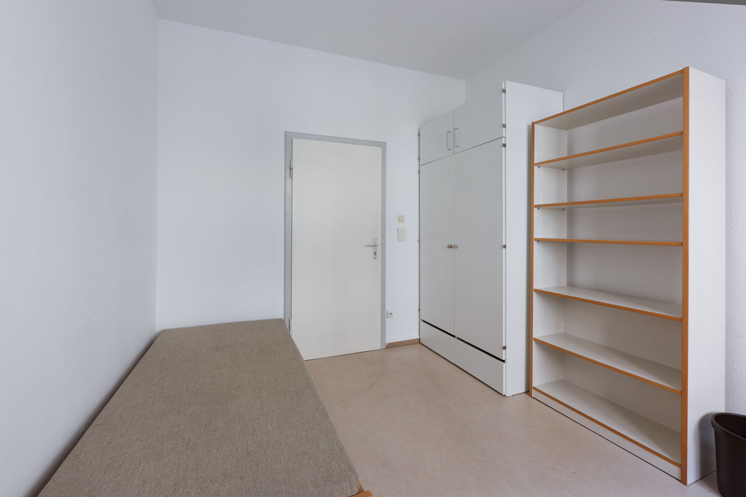 Schlicht eingerichtetes Zimmer mit hellem Boden, einem Einzelbett mit grauem Bezug, einem weißen Kleiderschrank, einem offenen Regal und einer weißen Tür; neutrale Wandfarbe und minimalistisches Design.