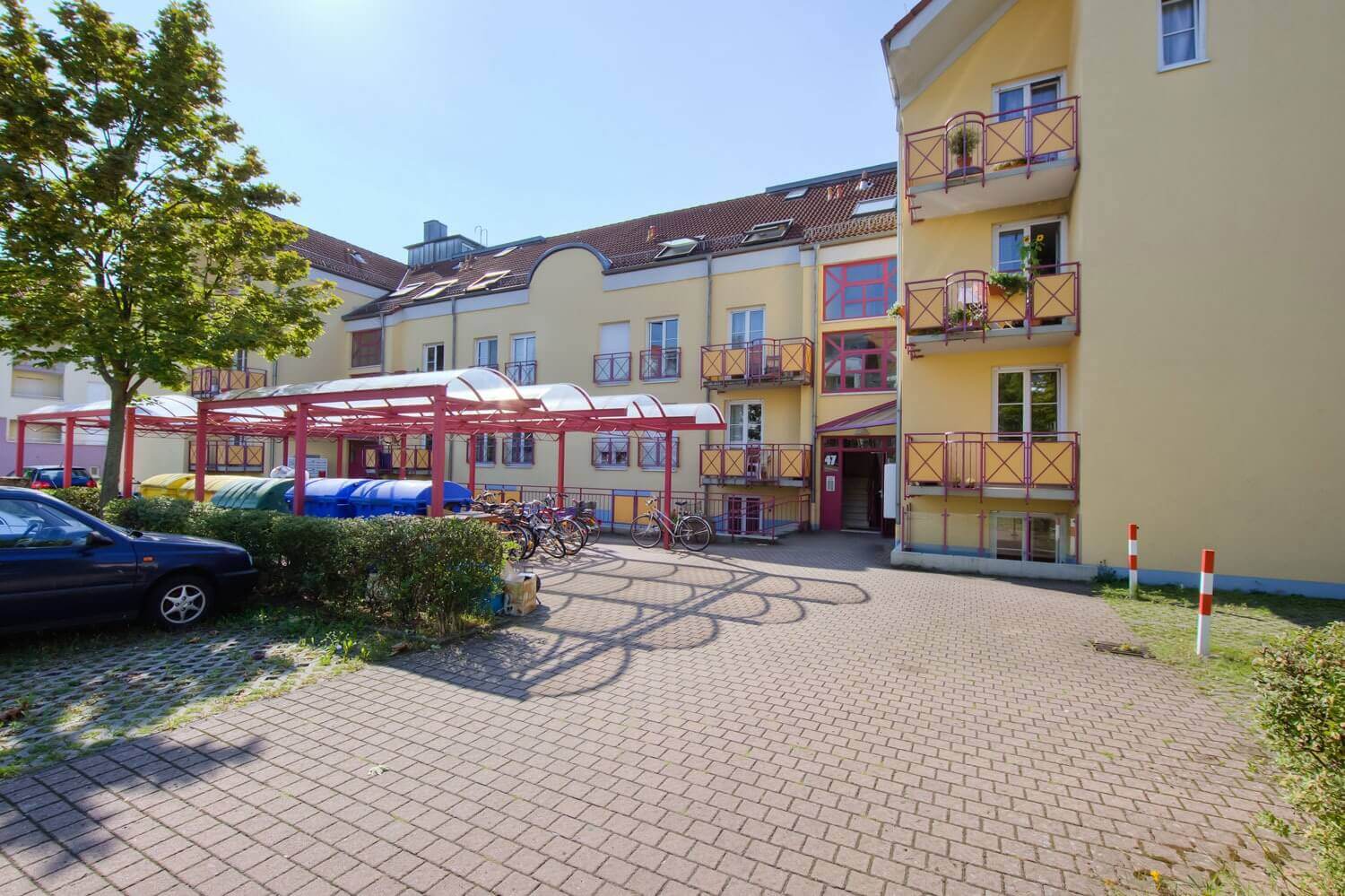 Courtyard side of a student residence with covered bicycle parking spaces, garbage containers, parked car and an entrance with house number 47