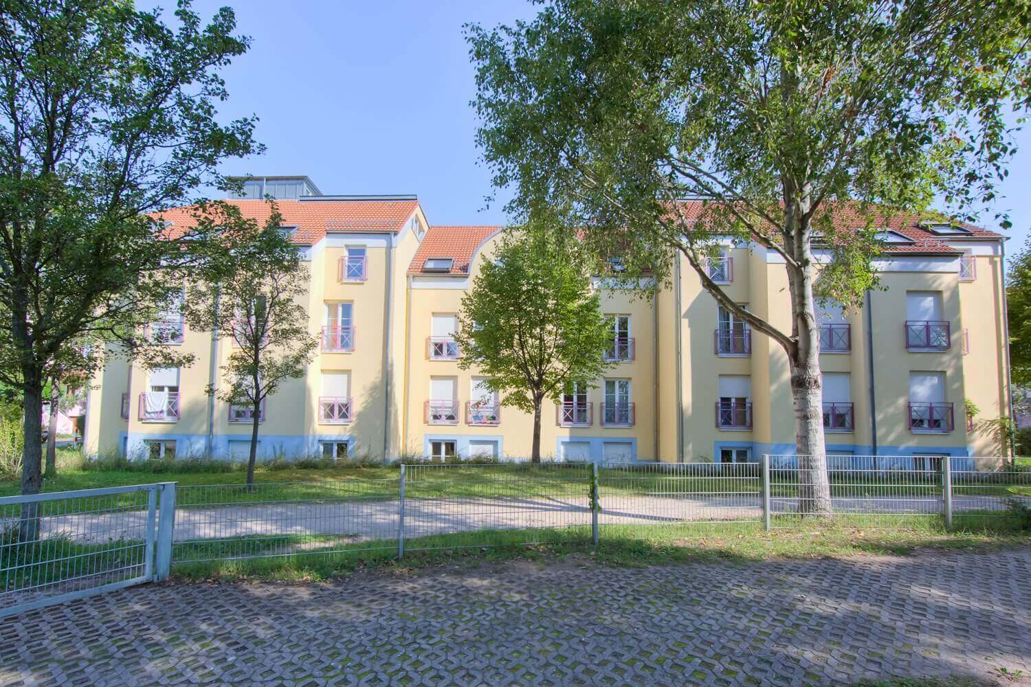 Yellow student residence with red window frames, red roof and small balconies, surrounded by trees and meadows