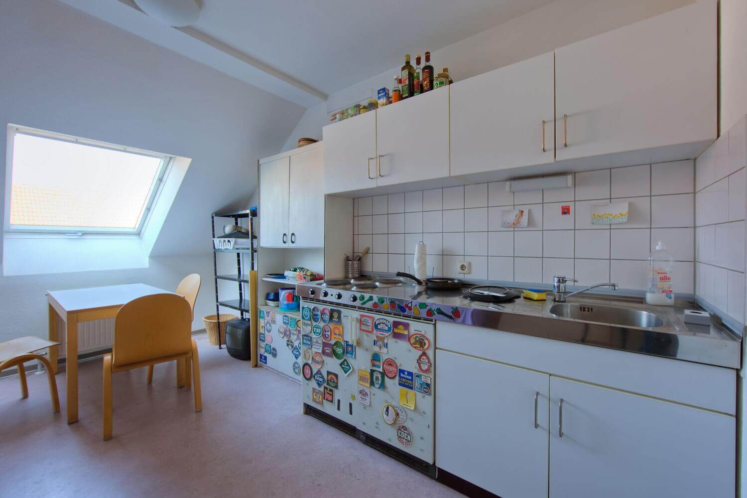 Shared kitchen with stove, sink and dining table under a sloping roof, the fridge is decorated with stickers