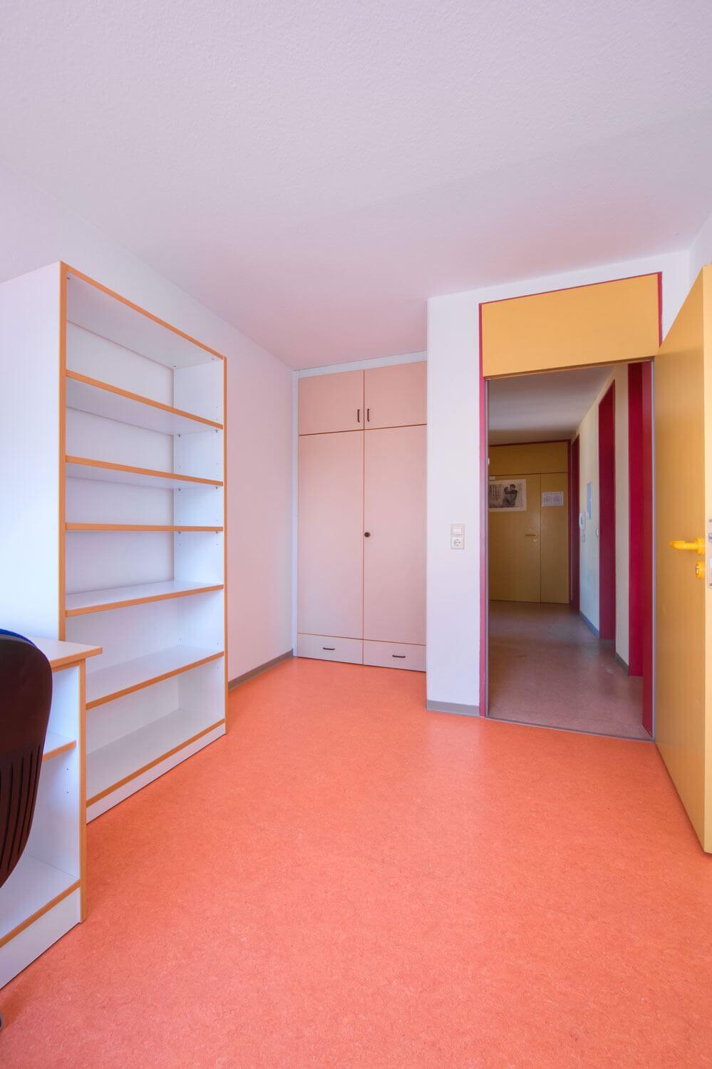 Single room in a student residence with bed, desk, shelf and view of a colorful hallway with built-in wardrobe