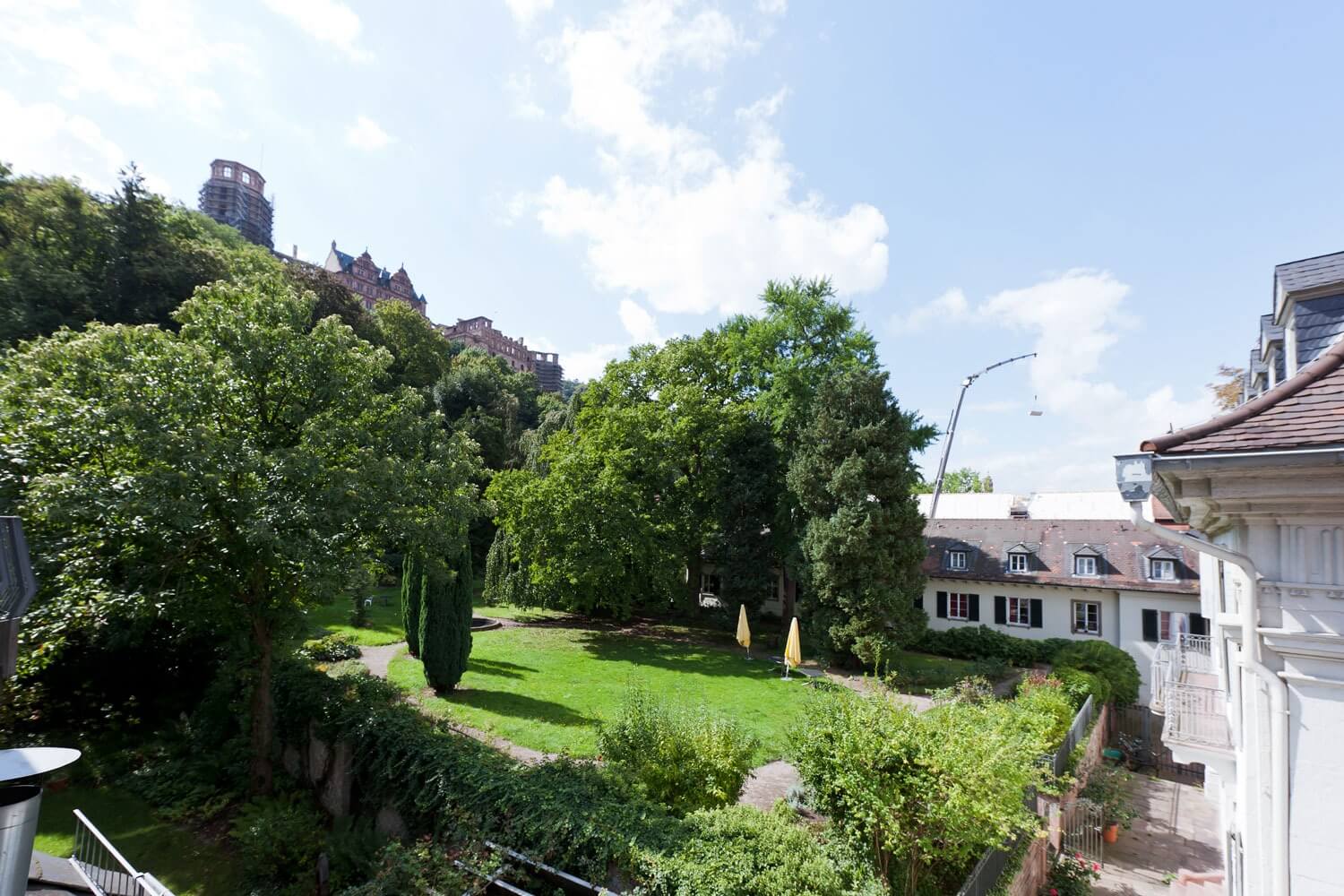 Ruhiger, begrünter Gartenbereich hinter dem Haus mit Rasen, Bäumen, Sonnenschirmen und Blick auf das Heidelberger Schloss, das auf einem bewaldeten Hügel thront.
