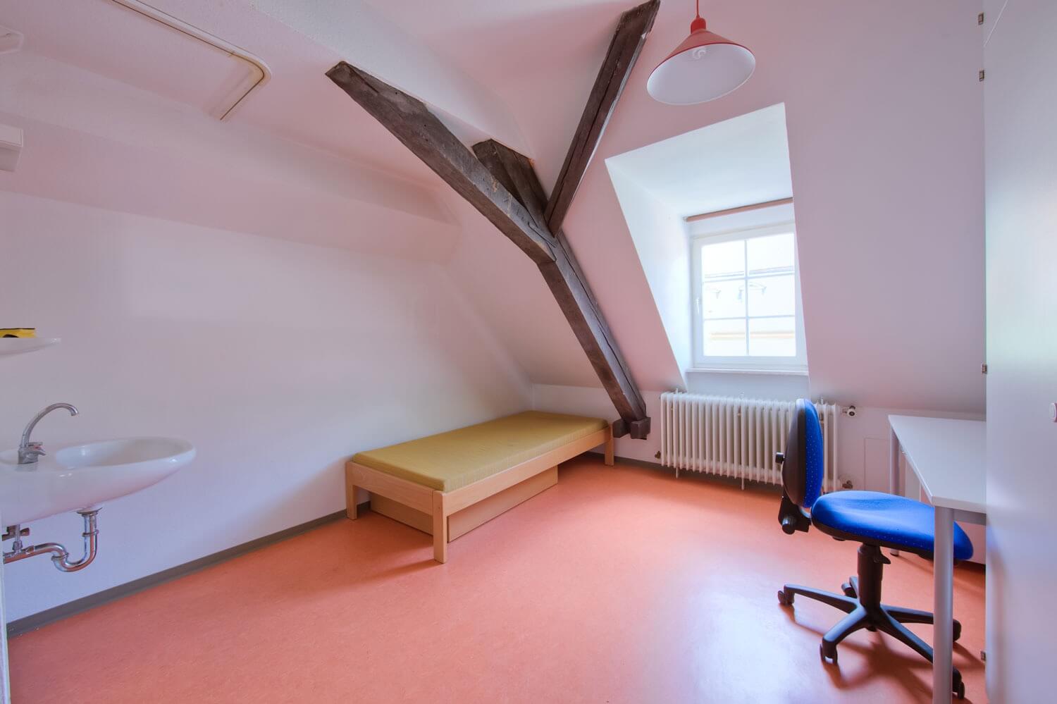 Gemütliches Zimmer im Dachgeschoss mit sichtbarem dunklem Holzbalken, Einzelbett, Schreibtisch mit Stuhl und einem Waschbecken; durch das Fenster fällt Tageslicht auf den orangefarbenen Boden.