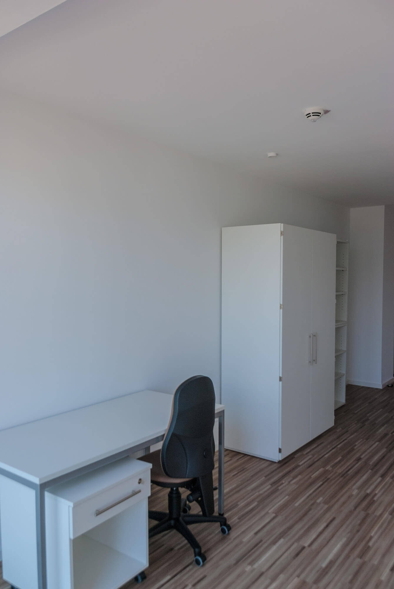 Long student room with desk, closet and shelves on a white wall, furnished in a simple, modern style.