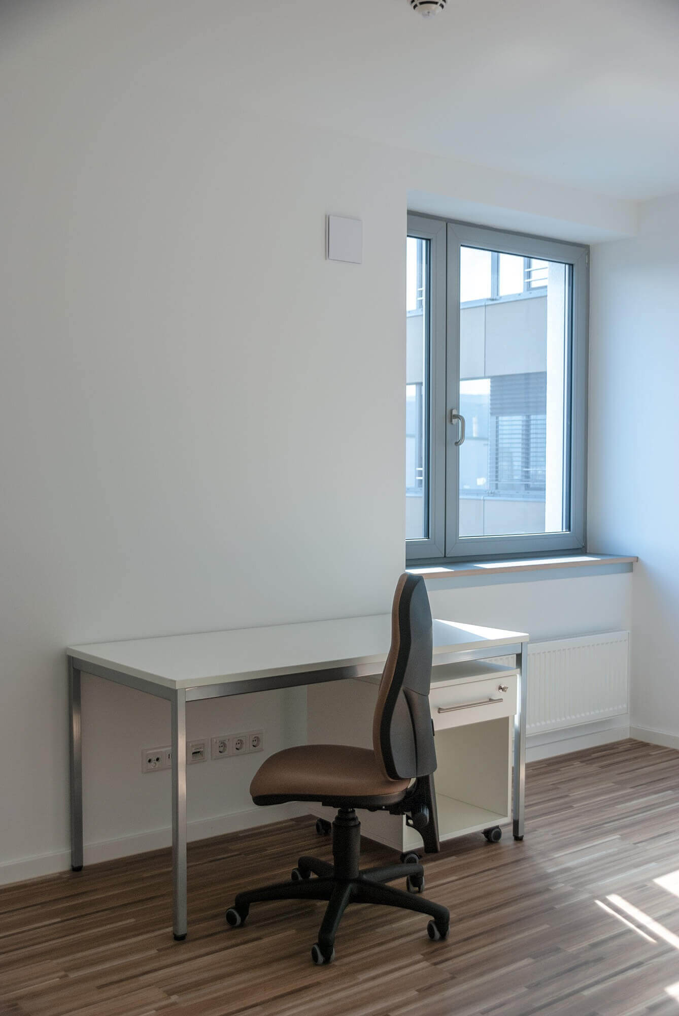 Bright workspace in the student residence with white desk, brown office chair and view of a large window.
