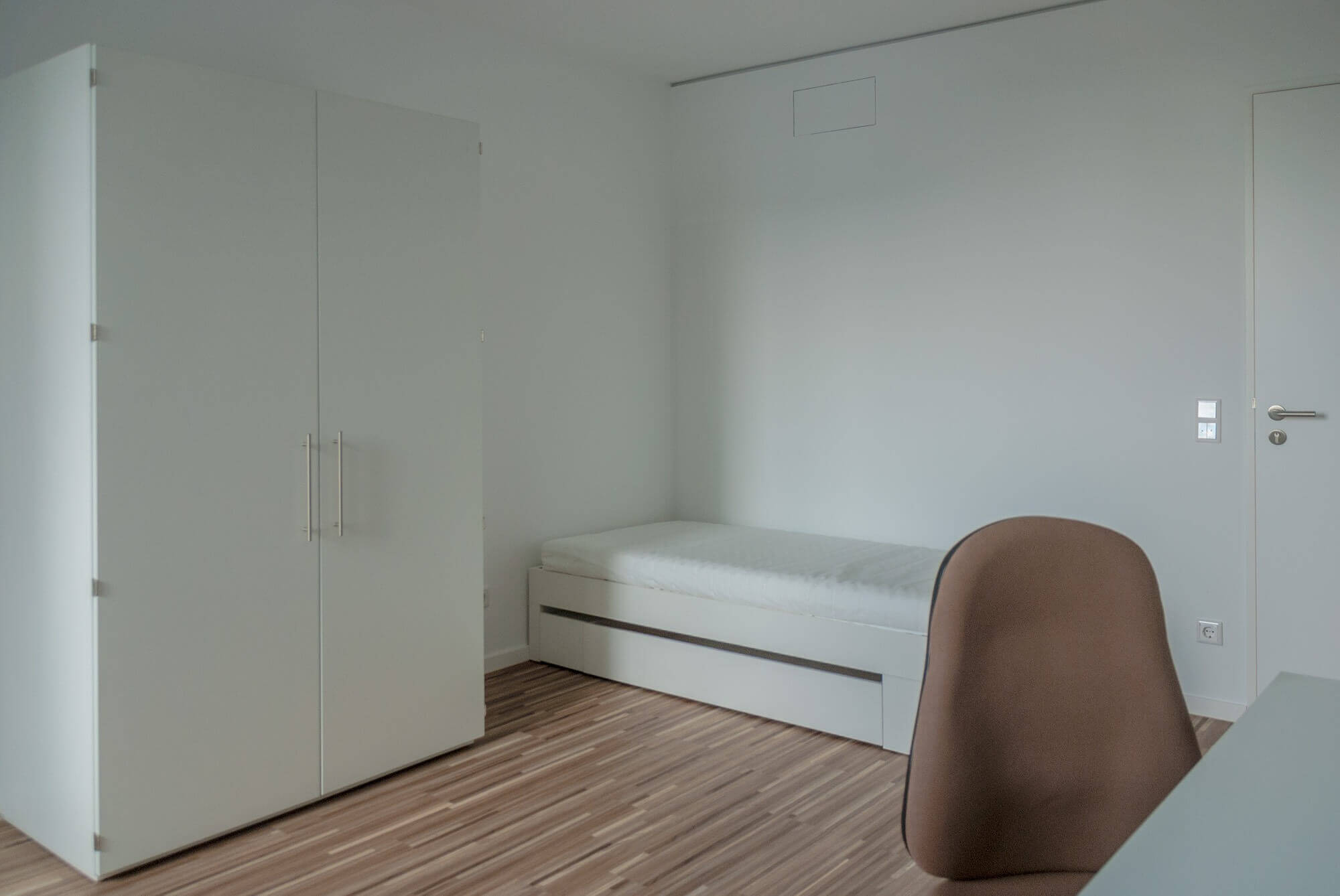 Simply furnished room in student residence with single bed, closet and desk chair on wooden floor.